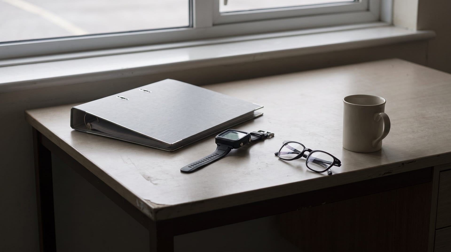 Carpeta de anillas cerrada sobre una mesa junto a una ventana, con un ordenador de buceo, unas gafas de leer y una taza al lado