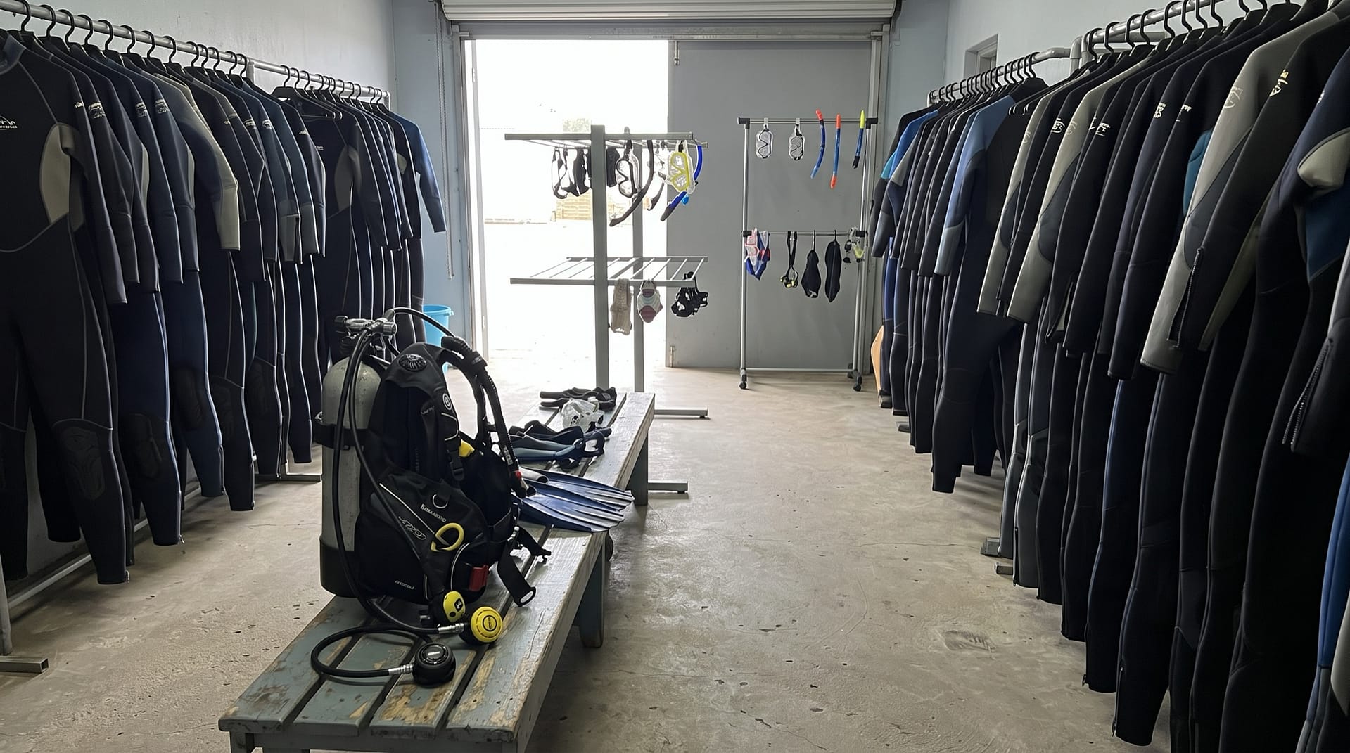 Sala de alquiler de un centro de buceo con dos hileras de trajes de neopreno colgados, máscaras y tubos en perchas, y un equipo montado con botella, jacket y regulador sobre un banco