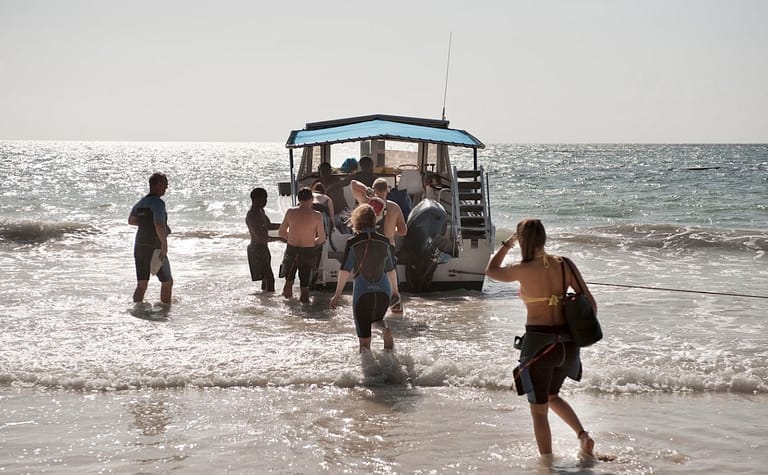 Grupo de buceadores con neopreno y equipo entrando al agua desde la playa para embarcar en una lancha de buceo en la costa de Kenia, a contraluz