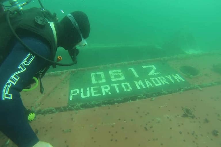 Buceador junto al casco de un pesquero hundido con la matrícula «0812 PUERTO MADRYN» pintada en la chapa, en el agua verde del Golfo Nuevo