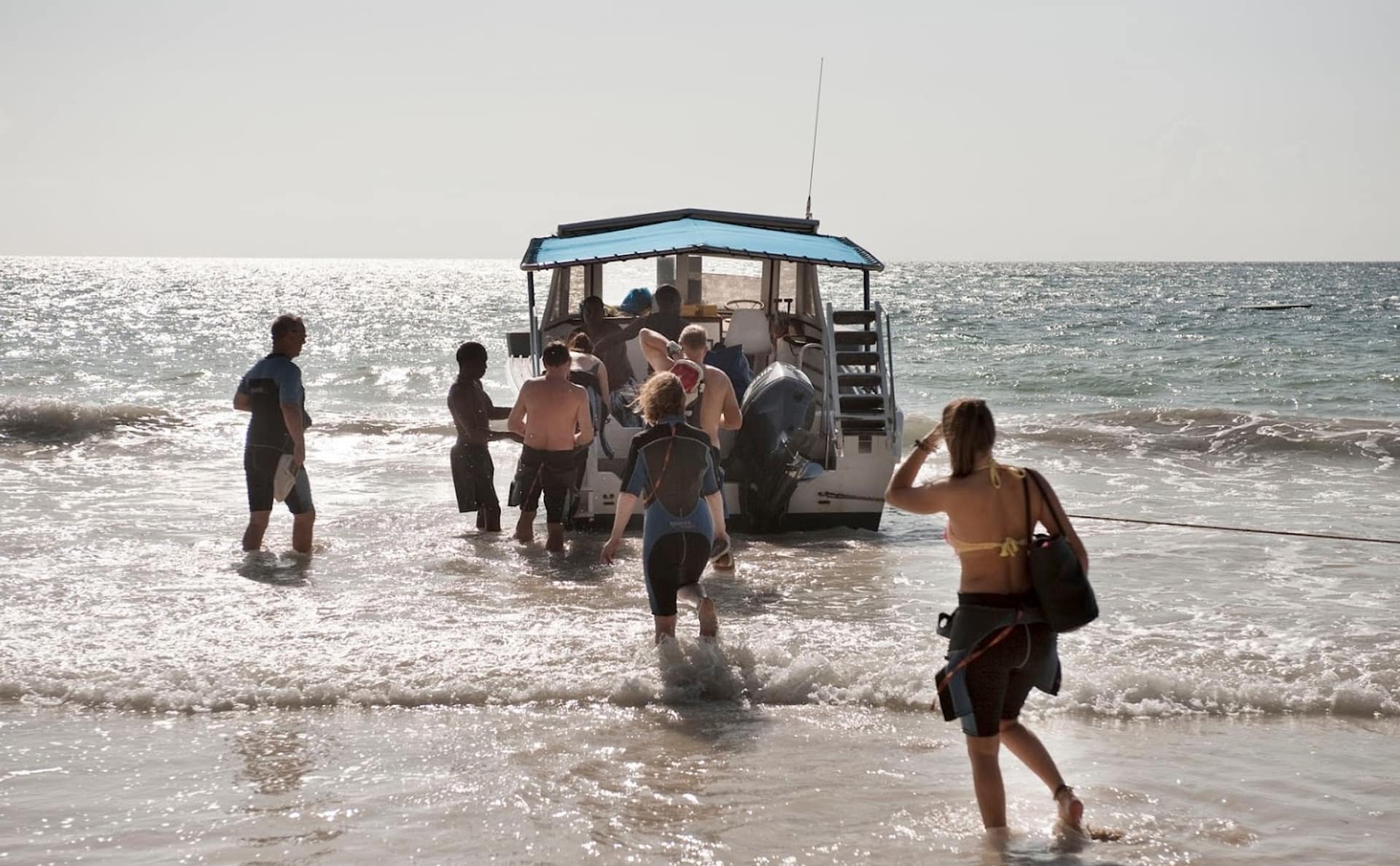 Grupo de buceadores con neopreno y equipo entrando al agua desde la playa para embarcar en una lancha de buceo en la costa de Kenia, a contraluz