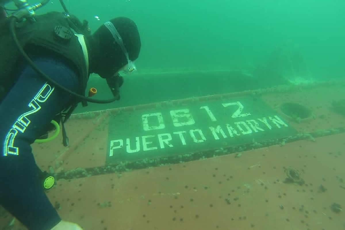 Buceador junto al casco de un pesquero hundido con la matrícula «0812 PUERTO MADRYN» pintada en la chapa, en el agua verde del Golfo Nuevo