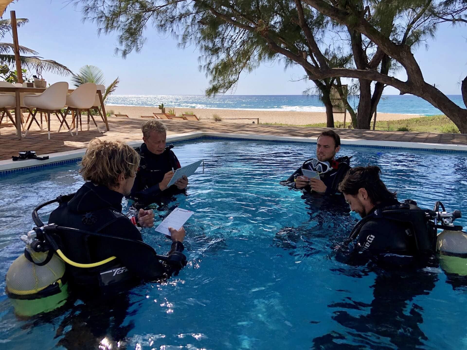 Instructor y tres alumnos con equipo completo dentro de una piscina de aguas confinadas, con pizarras en la mano y el mar al fondo